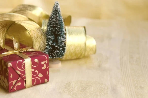 Christmas decoration: red gift box, gold ribbon and mini fir tree on golden background. Winter holiday. Christmas or  New Year concept. Copy space