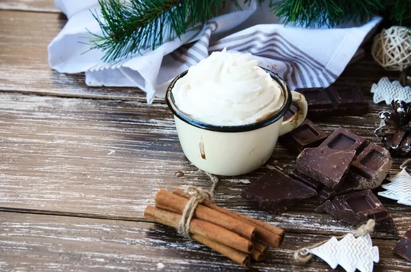 Enamel mug of hot chocolate cocoa with whipped cream, slice of bitter chocolate and cinnamon sticks on wooden vintage background. Winter times drink concept. Delicious cold weather beverage.
