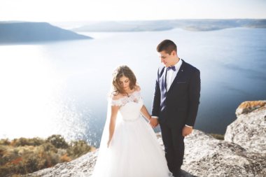 Düğün çifti mavi denizin yanındaki kayalarda öpüşüyor ve sarılıyor.