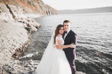 Düğün çifti mavi denizin yanındaki kayalarda öpüşüyor ve sarılıyor.