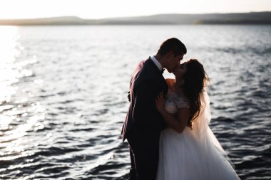 Düğün çifti mavi denizin yanındaki kayalarda öpüşüyor ve sarılıyor.