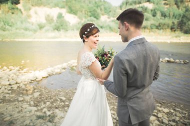 Güzel düğün çifti bir dağ nehrinin kıyısında taşlarla öpüşüp kucaklaşıyorlar.