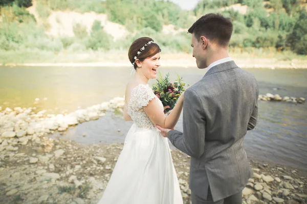 Güzel düğün çifti bir dağ nehrinin kıyısında taşlarla öpüşüp kucaklaşıyorlar.