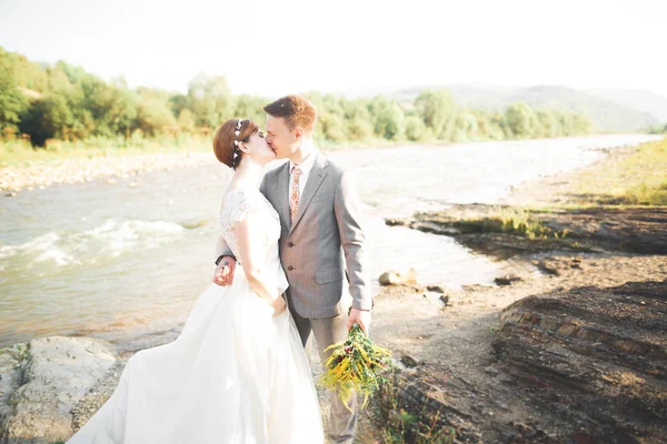 Güzel düğün çifti bir dağ nehrinin kıyısında taşlarla öpüşüp kucaklaşıyorlar.