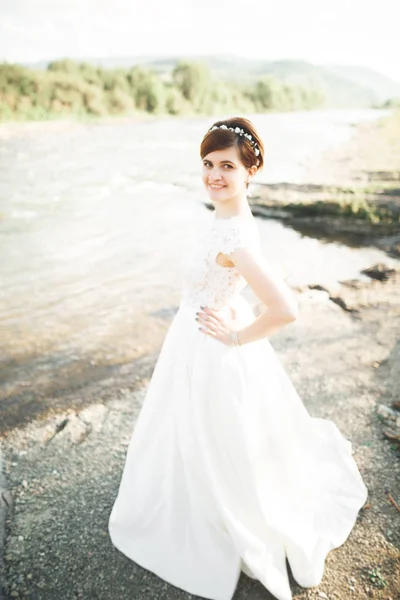 Splendida bella sposa sorride mentre posa sul fiume con bouquet — Foto Stock