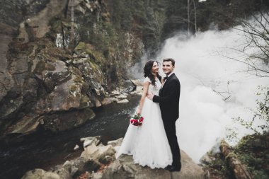 Birbirlerine aşık düğün çifti güzel manzarada kayaların yanında öpüşüyor ve sarılıyorlar.