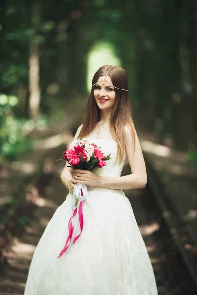 Bella sposa bruna in elegante vestito bianco che tiene bouquet in posa alberi ordinati — Foto Stock