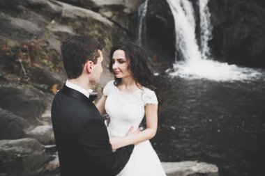 Birbirlerine aşık düğün çifti güzel manzarada kayaların yanında öpüşüyor ve sarılıyorlar.