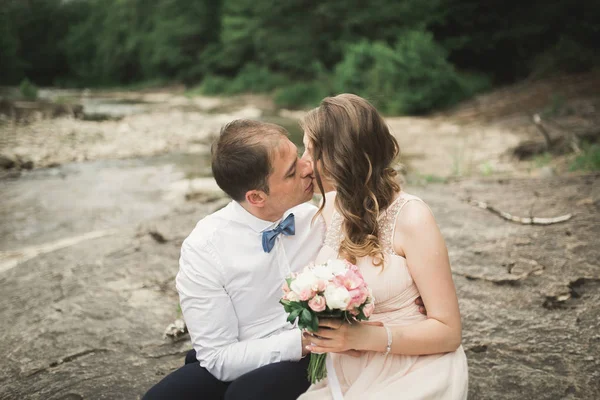 Beautifull wedding couple kissing and embracing near the shore of a mountain river with stones — Stock Photo, Image