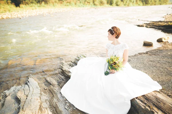 Splendida bella sposa sorride mentre posa sul fiume con bouquet — Foto Stock