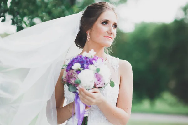 Красивая невеста в элегантном белом платье держа букет позируя в парке — стоковое фото