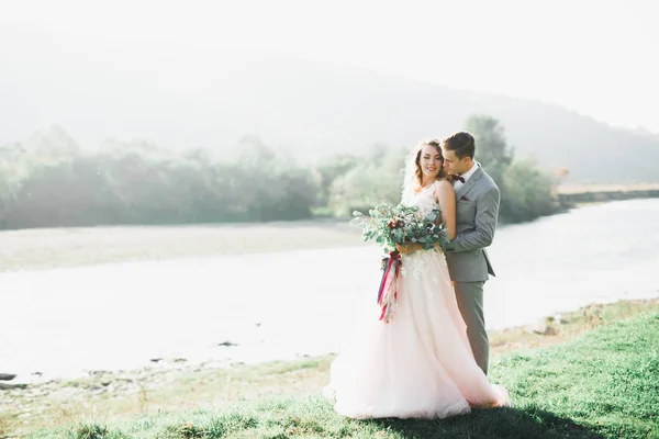 Romantisches, märchenhaftes, glückliches Brautpaar, das sich im Park umarmt und küsst, Bäume und Fluss im Hintergrund — Stockfoto
