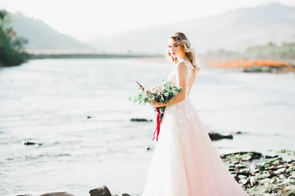 Krásná brunetka nevěsta v elegantních bílých šatech držení kytice pózuje v blízkosti řeky — Stock fotografie