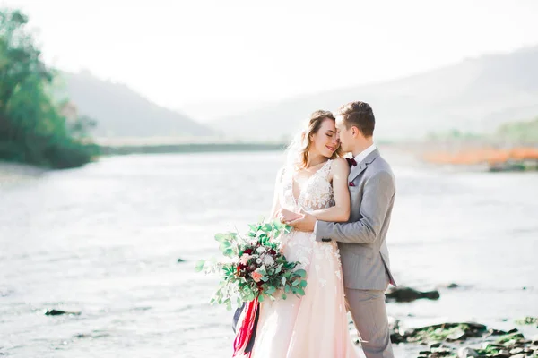 Romantic, fairytale, happy newlywed couple hugging and kissing with trees and river in background — Stock Photo, Image