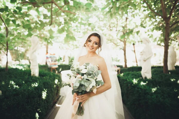 Belle mariée brune en robe blanche élégante tenant bouquet posant des arbres soignés — Photo