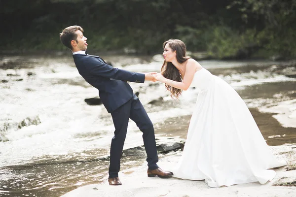 Hermosa pareja de boda besándose y abrazándose cerca de la orilla de un río de montaña con piedras —  Fotos de Stock