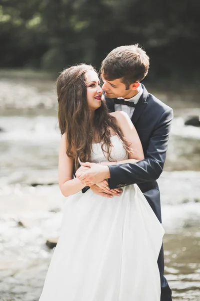 Elegante elegante novia morena feliz y hermoso novio en el fondo de un hermoso río en las montañas —  Fotos de Stock