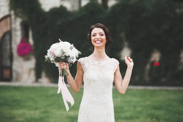 Hermosa novia morena en elegante vestido blanco sosteniendo ramo posando árboles limpios — Foto de Stock