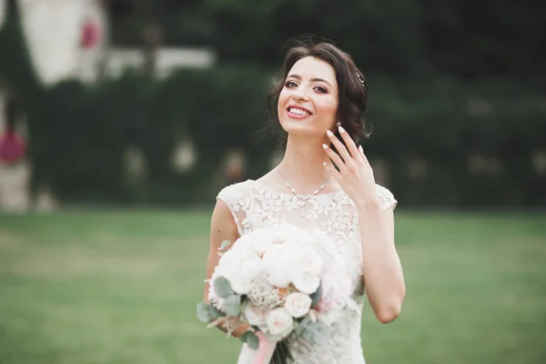 Bella sposa bruna in elegante vestito bianco che tiene bouquet in posa alberi ordinati — Foto Stock