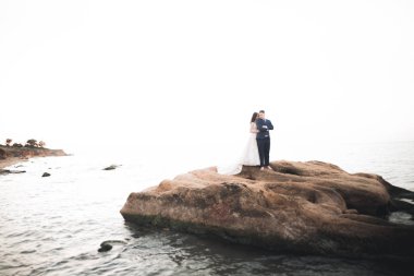 Düğün çifti mavi denizin yanındaki kayalarda öpüşüyor ve sarılıyor.