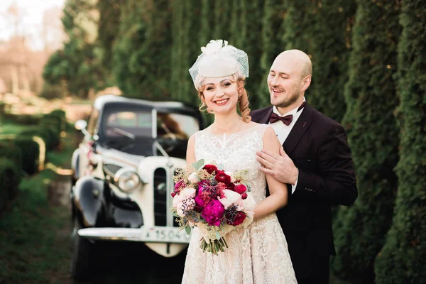Feliz casal recém-casado, homem e mulher beijando perto de carro retro elegante — Fotografia de Stock