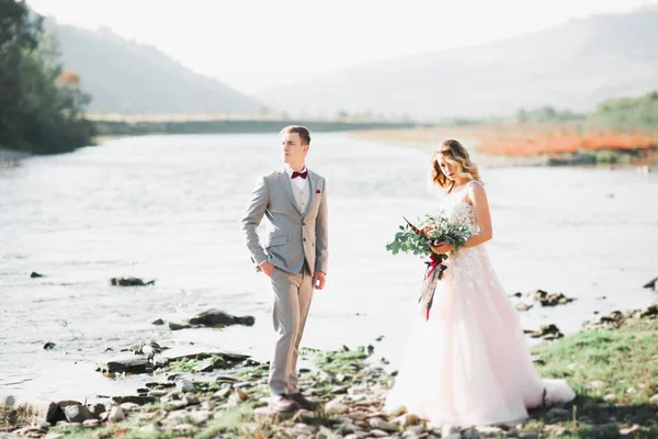 Elegante novia rubia feliz y elegante novio en el fondo de un hermoso río en las montañas —  Fotos de Stock