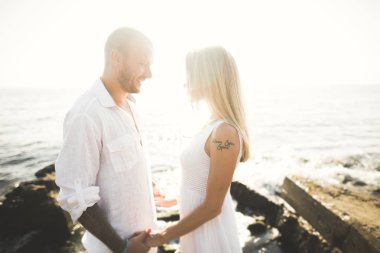 Düğün çifti mavi denizin yanındaki kayalarda öpüşüyor ve sarılıyor.