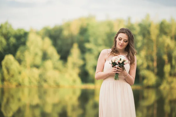 Linda noiva em vestido elegante segurando buquê posando perto da floresta e do lago — Fotografia de Stock