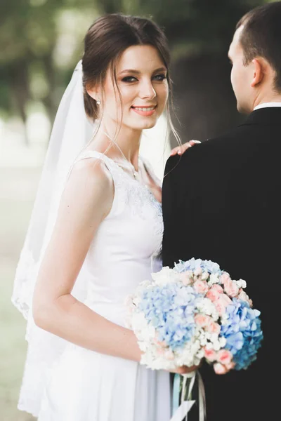 Elegante coppia di sposi felici che passeggiano nel parco il giorno del loro matrimonio con bouquet — Foto Stock