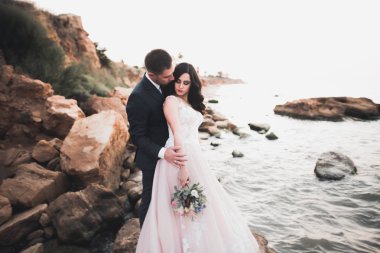 Düğün çifti mavi denizin yanındaki kayalarda öpüşüyor ve sarılıyor.