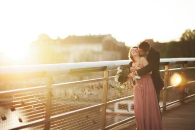 Şık Düğün çifti, damat, Gelin öpüşme ve günbatımında bir köprüde sarılma pembe elbise ile sevgi dolu