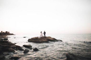 Düğün çifti mavi denizin yanındaki kayalarda öpüşüyor ve sarılıyor.