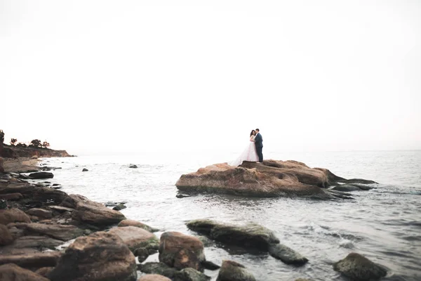 Düğün çifti mavi denizin yanındaki kayalarda öpüşüyor ve sarılıyor. — Stok fotoğraf