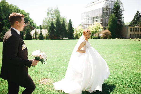 Stilvolles Paar glücklicher Frischvermählter. Braut läuft dem Bräutigam am Hochzeitstag mit Blumenstrauß im Park davon — Stockfoto