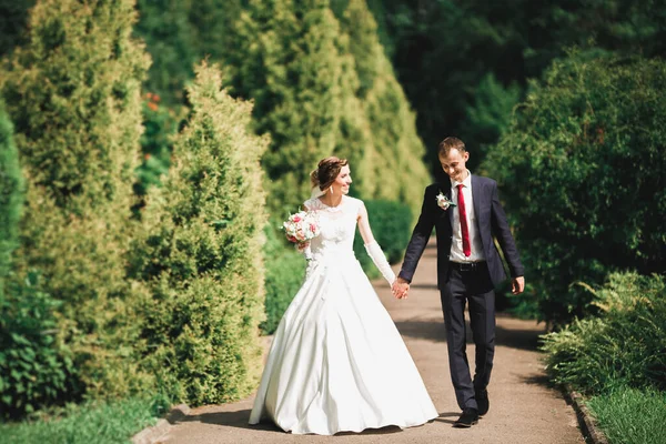 Κομψό ζευγάρι χαρούμενων νεόνυμφων που περπατούν στο πάρκο την ημέρα του γάμου τους με μπουκέτο — Φωτογραφία Αρχείου