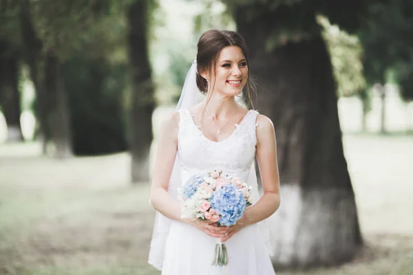 Hermosa novia en elegante vestido blanco sosteniendo ramo posando en el parque — Foto de Stock