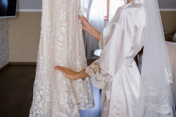 A noiva em um roupão de banho fica perto do vestido de casamento — Fotografia de Stock