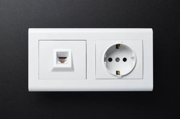 An electric wall socket and a white telephone socket are located next to a black background.