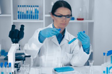 Beyaz önlüklü kendini adamış bir kadın bilim adamı, laboratuvar ekipmanlarıyla çevrili bir deney tüpünü ve pipetini ustalıkla kullanıyor, titizlikle kimyasal reaksiyonları inceliyor ve dikkatle inceliyor.