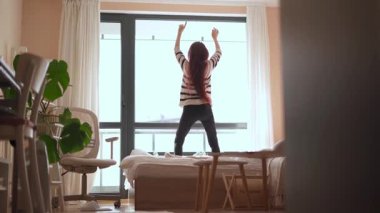 Canlı, genç bir kadın rahat yatağında neşe içinde dans eder, şık, modern yaşam alanını dolduran güzel sabah ışığını kucaklar, ki bu da her yerde canlı, yemyeşil bitkilerle süslenir.