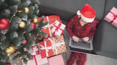 Noel Baba şapkalı ve renkli pijamalı neşeli bir çocuk dizüstü bilgisayarında, etrafı süslenmiş bir ağacın altında parıldayan ışıklarla çevrili, neşeli bir tatil sahnesi yaratıyor.