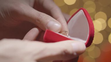 Yakından bakınca, kadife bir kırmızı yüzük kutusunu incelikle açan, içinde çarpıcı bir nişan yüzüğü olan, sahte bir arkaplan oluşturan, aşkın ve bağlılığın özünü yakalayan eller görülüyor..