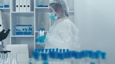 Cam eşyalar ve deneysel tertibatlarla donatılmış modern bir laboratuvarda, koruyucu giysili kendini adamış bir bilim adamı örnekleri inceleyip araştırma için yüksek teknolojili bir mikroskop kullanıyor.