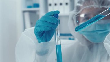 Steril bir laboratuvarda, koruyucu giysiler içindeki odaklanmış bir bilim adamı deney tüplerini parlak mavi çözeltilerle inceleyerek kritik deneyler sırasında olağanüstü hassasiyet ve detaya dikkat gösteriyor.