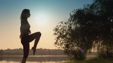 Şafak vakti, bir kadın dingin bir gölün kenarında yoga yapıyor, silueti doğayla uyum ve farkındalığı somutlaştırıyor, rahatlama ve refah telkin eden bu huzur ortamında sükunet yayıyor.