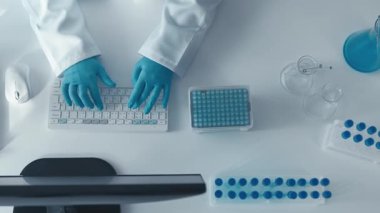 Beyaz önlüklü bir bilim adamı, laboratuvar araştırmalarında hassasiyet ve yeniliğin önemini vurgulayan klavye, pipet ve gelişmiş laboratuvar aletleriyle verileri analiz eder.