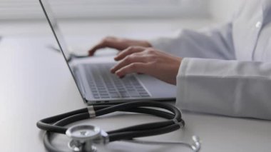 Beyaz önlüklü bir sağlık uzmanı hasta bilgilerini bir dizüstü bilgisayara, masanın üzerinde steteskop ile yerleştiriyor. Hasta bakımının iyileştirilmesi için teknoloji ve sağlık hizmetlerinin kesişimini gösteriyor.