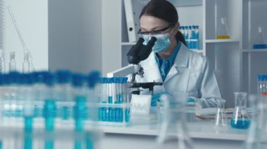 Beyaz laboratuvar önlüklü kendini adamış bir bilim adamı ileri laboratuvar ekipmanlarıyla çevrili örnekleri mikroskop altında inceliyor ve araştırmada hassasiyet ve doğruluğun önemini vurguluyor.