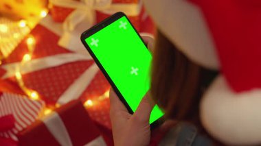 Samimi bir tatil sahnesinde Noel Baba şapkalı bir kişi, yeşil ekranlı akıllı bir telefonla neşeli Noel hediyeleriyle mutlu bir şekilde geziyor ve neşeli atmosfere neşe katıyor.