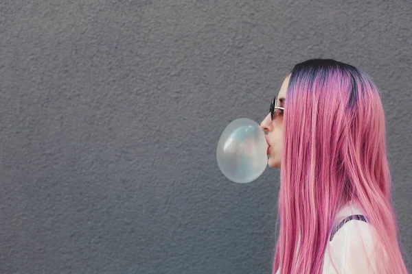 Elegante jovem hipster mulher com cabelo rosa longo soprando uma bolha com chiclete . — Fotografia de Stock